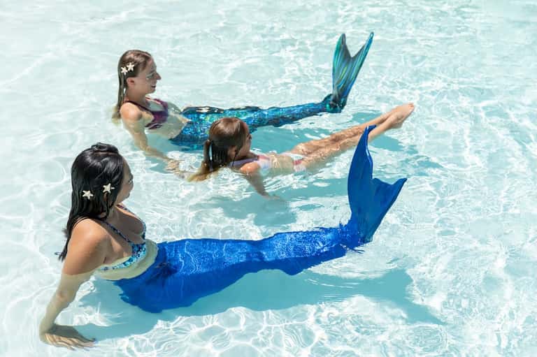 Children in colorful mermaid tails swimming in shallow turquoise resort pool water