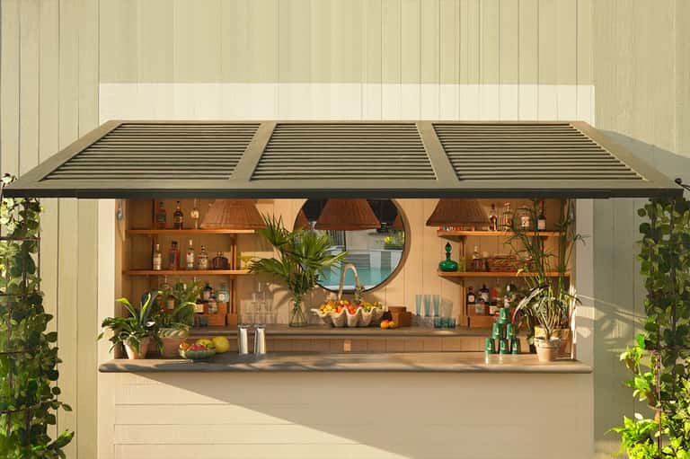 Outdoor bar counter with wooden shelving, bottles, plants, round mirror, pendant lights, and retractable awning