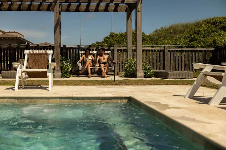 Two guests on porch swing overlooking small pool with wooden pergola and beach landscaping