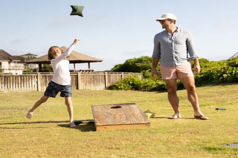 Father and child playing cornhole on resort lawn with wooden buildings and palm trees in background
