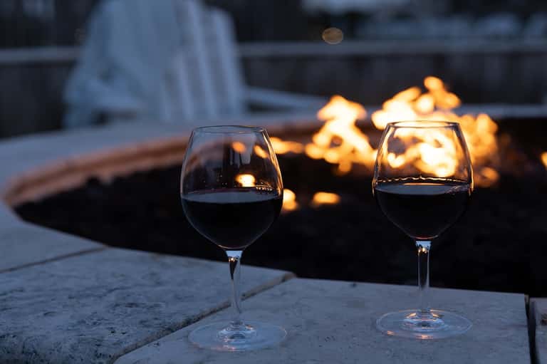 Two wine glasses on stone surface with glowing fire pit and outdoor seating in evening light