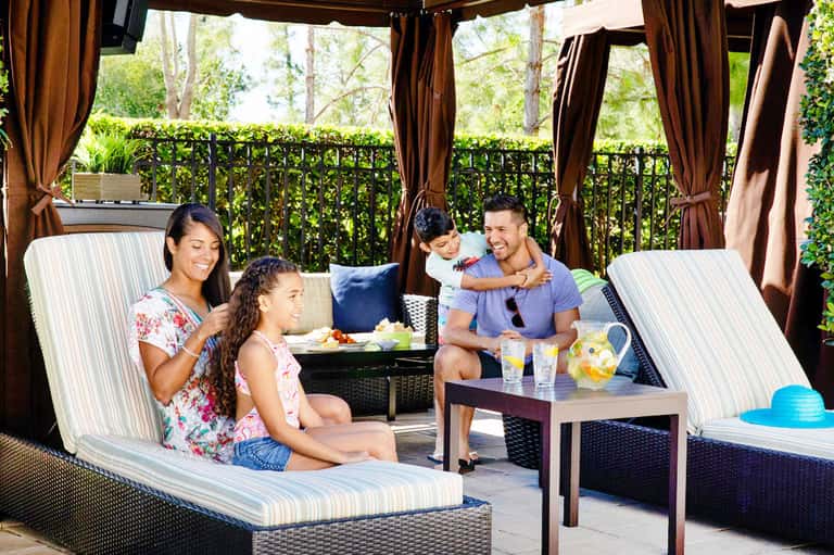 Family of four relaxing on lounge chairs under brown cabana with striped cushions and curtains