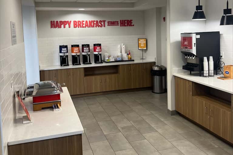 Hotel breakfast area with coffee dispensers, signage reading 'Happy Breakfast Starts Here'