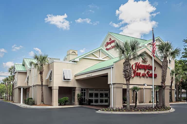 Beige hotel building with green metal roof, palm tree landscaping, and American flag in sunny Florida setting