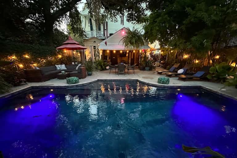 Backyard pool illuminated with blue lights at dusk, surrounded by lounge seating and trees