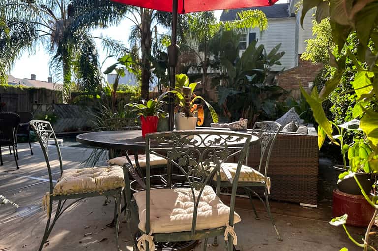 Outdoor patio with round table, metal chairs, red umbrella, tropical plants, and white house structure
