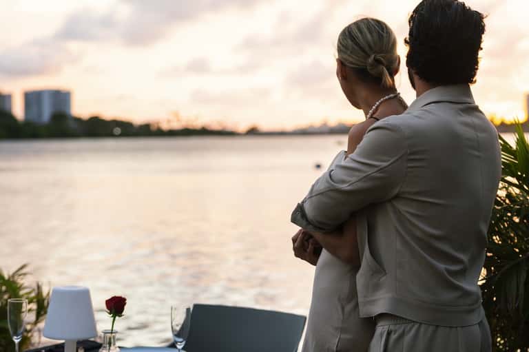 Couple embracing on waterfront balcony at sunset with city skyline reflected in water
