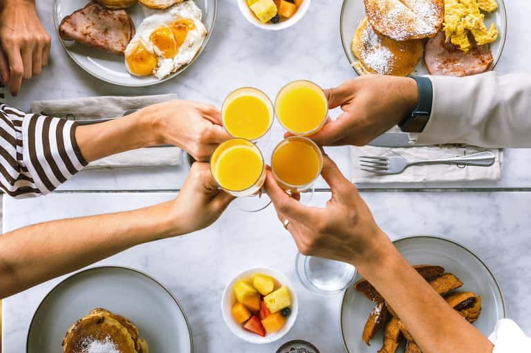 Overhead view of brunch table with four hands holding glasses of orange juice, pastries, eggs, and fresh fruit