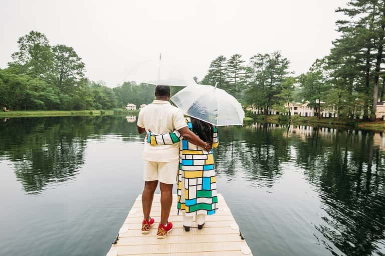 Couple with umbrella standing on dock overlooking calm lake surrounded by lush green trees and homes