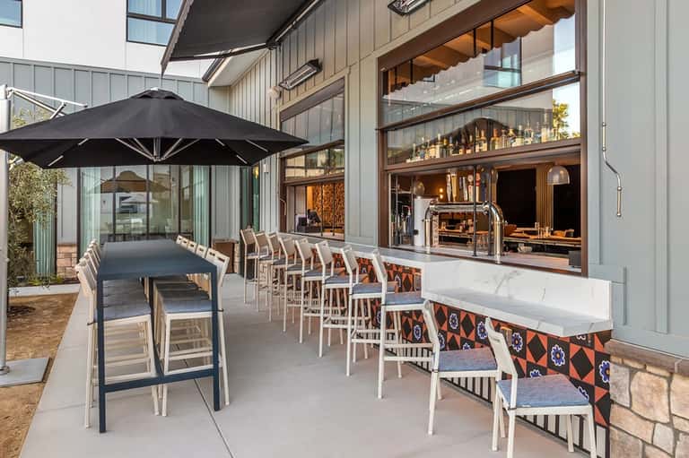 Modern patio dining area with long table, black umbrella, white chairs, and full-service outdoor kitchen