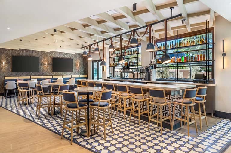 Contemporary hotel bar with wooden chairs, geometric patterned rug, and fully stocked shelving