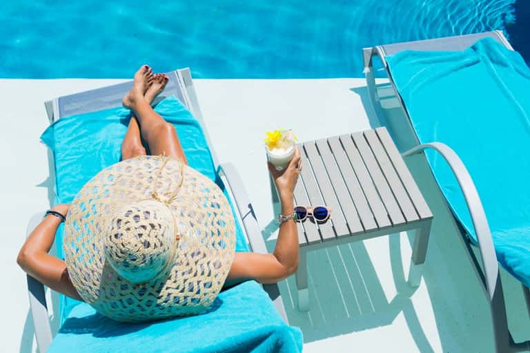 Overhead view of resort guest lounging by bright blue pool with hat and tropical cocktail