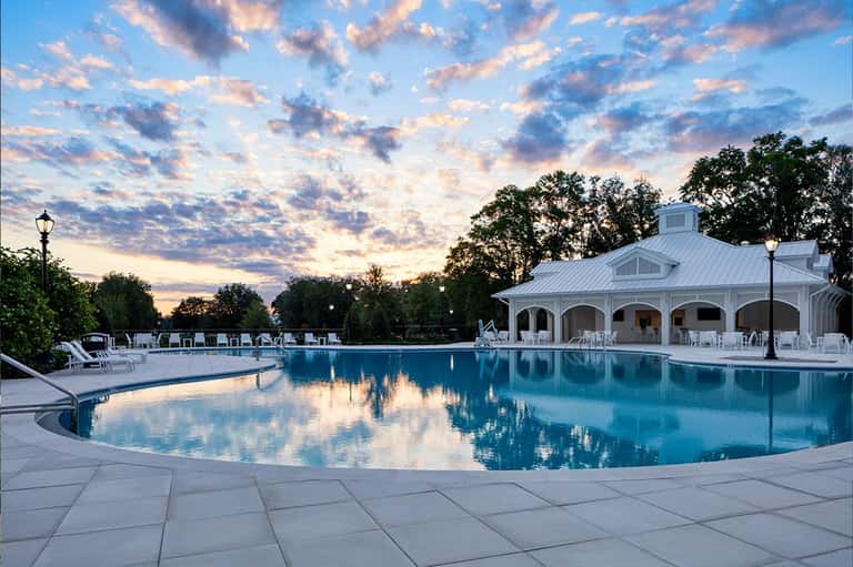 Elegant resort pool at dusk with illuminated pool house, white lounge chairs, and sunset sky reflections