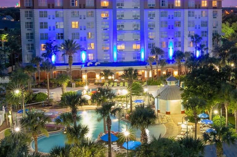 Nighttime aerial view of illuminated resort with blue-lit building, pool, palm trees, and landscaped grounds