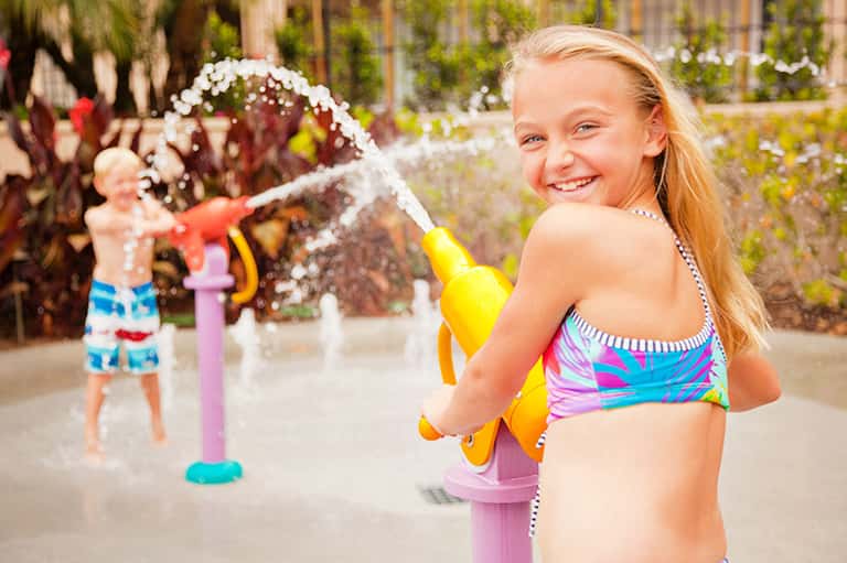 Children playing in a splash pad with water fountains and tropical plants in background