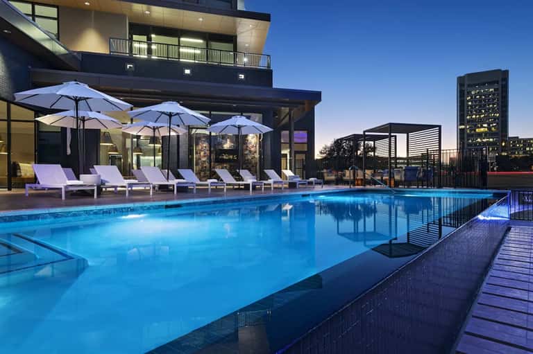 Rooftop swimming pool at dusk with lounge chairs, blue umbrellas, and city skyline backdrop