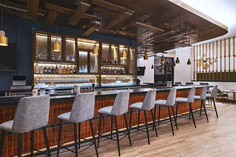 Modern bar with wooden accents, illuminated shelving, brass pendants, and bar seating