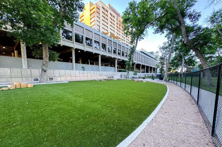 Urban dog park with synthetic grass, fencing, modern building structure, and surrounding trees