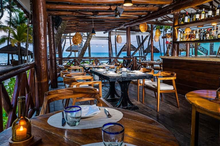 Rustic beachfront restaurant with wooden beams, tables set for dining, ocean and palm trees visible