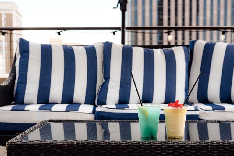 Two tropical cocktails on outdoor table with blue and white striped cushions and city skyline