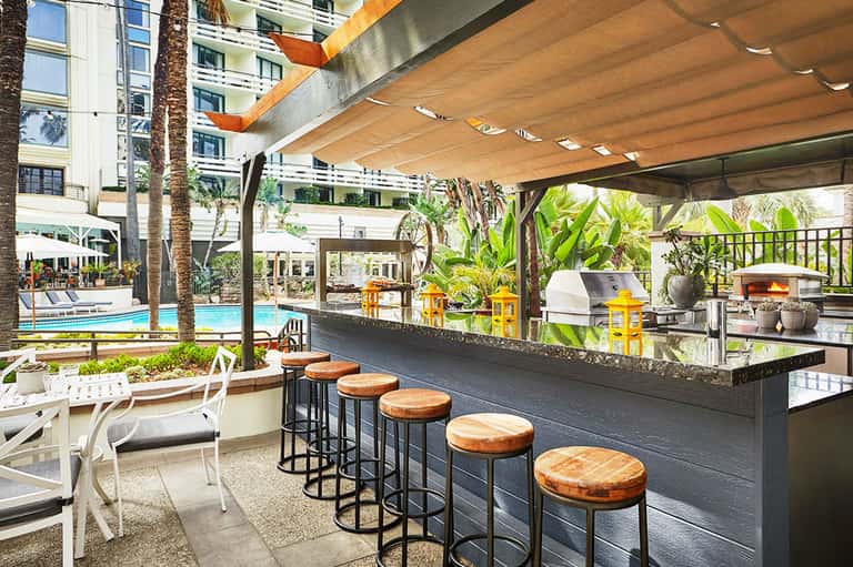Outdoor pool bar with wooden stools under a wooden pergola, palm trees, and resort buildings in background