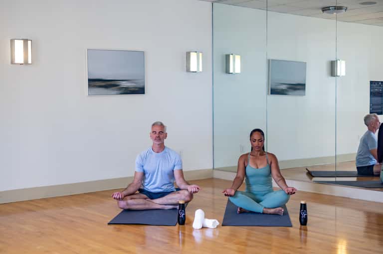 Yoga studio with two people meditating on mats, mirror wall, wooden floor, and minimalist coastal décor