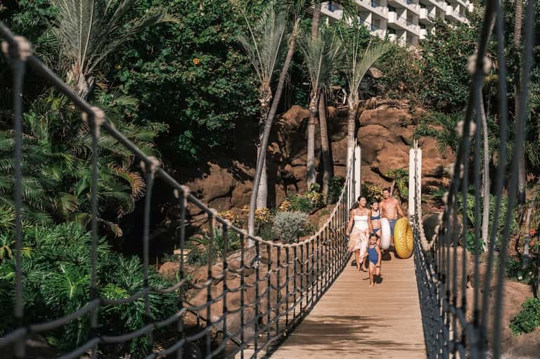 Family on wooden bridge over rocky terrain in lush tropical setting with palm trees and resort building
