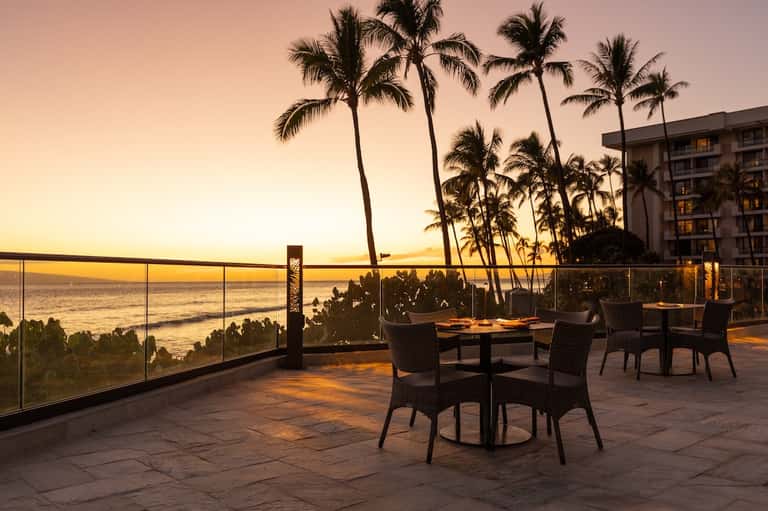 Oceanfront terrace with dining tables and palm trees at golden sunset