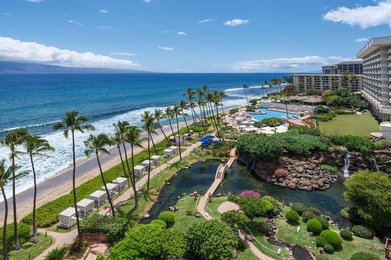 Aerial beachfront resort view with ocean, palm trees, pools, and manicured gardens
