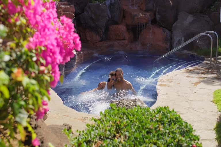 Two guests enjoying a hot spring pool surrounded by pink flowering plants and rocky landscape
