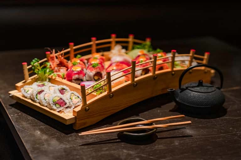 Artistic sushi platter arrangement in wooden boat with black teapot on dark surface
