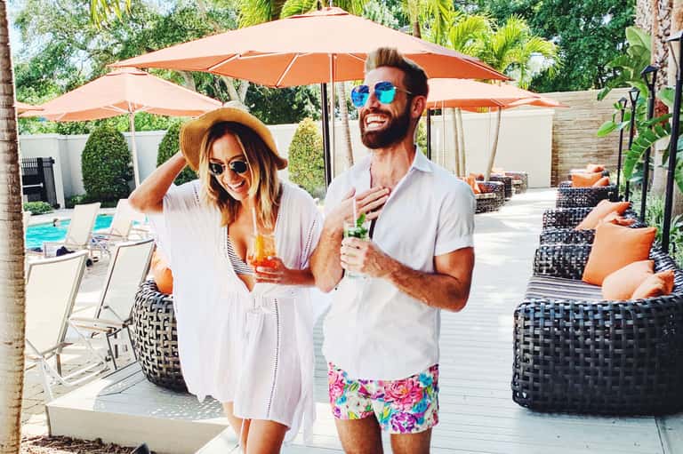 Couple enjoying drinks by resort pool under orange umbrellas with lounge chairs and palm trees