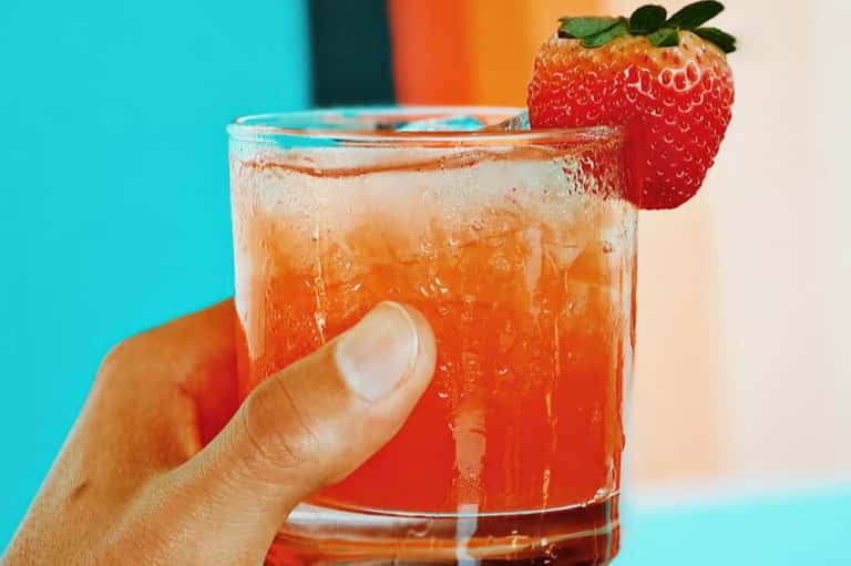 Strawberry-garnished cocktail with orange liquid and ice in vibrant turquoise setting