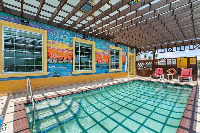 Indoor heated pool with colorful mural, yellow trim, wooden pergola ceiling, and lounge chairs poolside