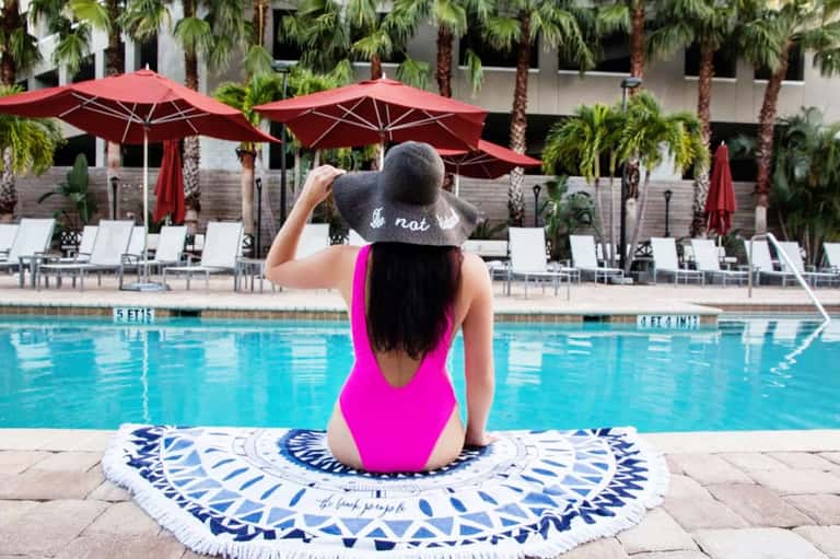 Woman in pink swimsuit sitting by resort pool with red umbrellas and palm trees in background