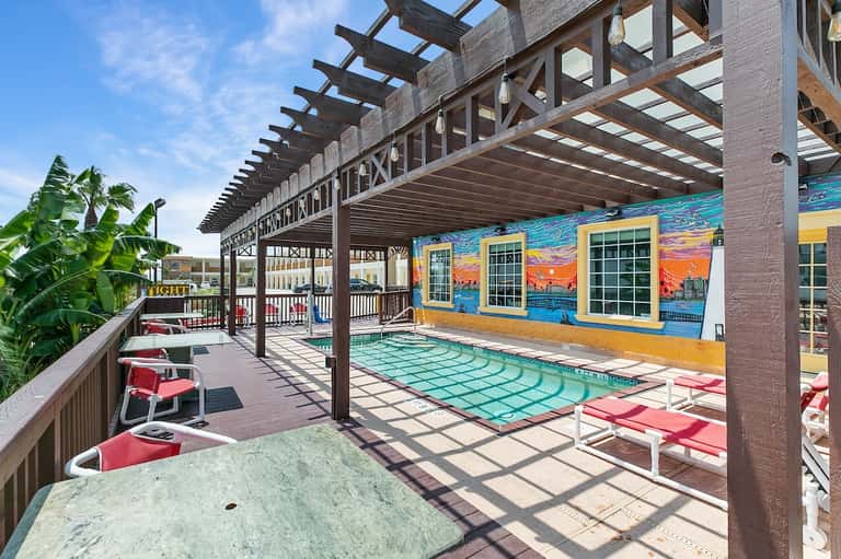 Covered pool deck with dark wooden pergola, turquoise water, colorful mural, and palm vegetation