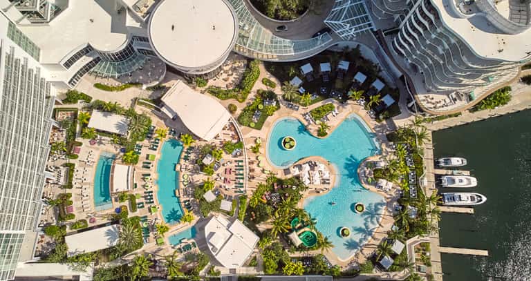 Aerial view of large resort pool complex with multiple pools, water features, palm trees, and surrounding buildings