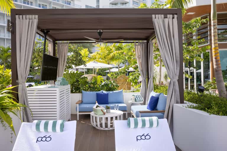 Outdoor lounge pavilion with gray curtains, blue cushioned seating, and tropical vegetation