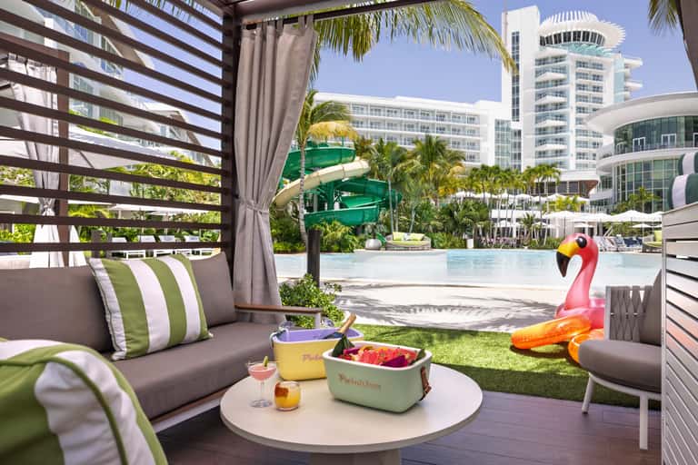 Tropical pool cabana with striped shade structure, water slide, and modern resort buildings