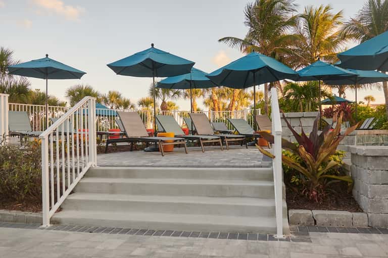 Beachside lounge area with blue umbrellas, palm trees, and colorful loungers