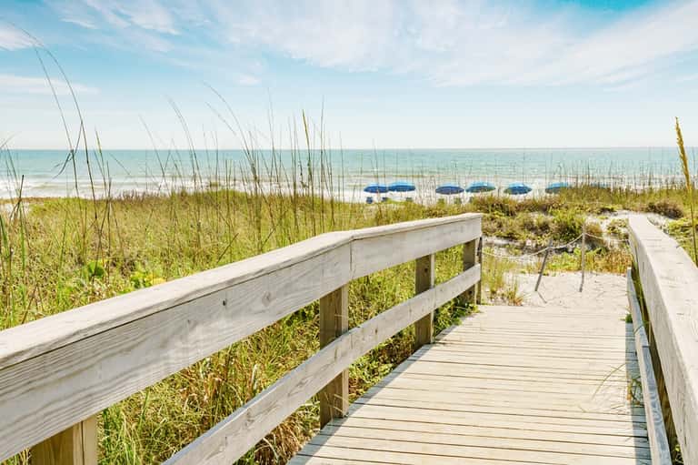 Wooden boardwalk leading to sandy beach with dune grass and blue umbrellas beyond the shoreline