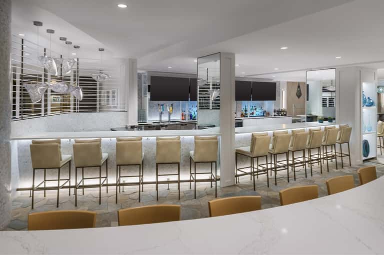 Modern hotel bar with marble countertops, gold bar stools, white pendant lighting, and mirrored walls