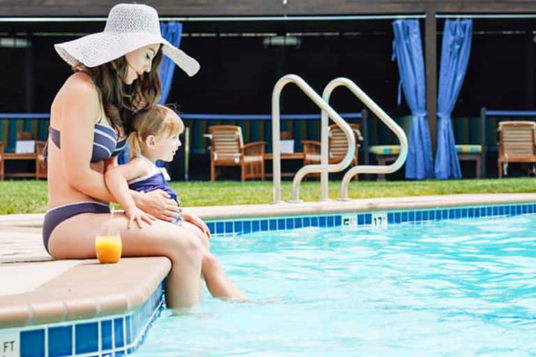 Woman and child sitting poolside wearing swimwear and sun hat by rectangular pool