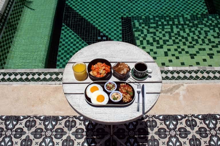 Breakfast tray by green tiled pool featuring eggs, fruit, pastries, juice, and coffee at waterside