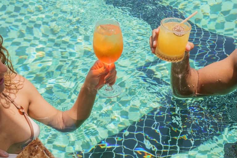 Guests holding colorful cocktails while swimming in clear turquoise pool with rippling water reflections