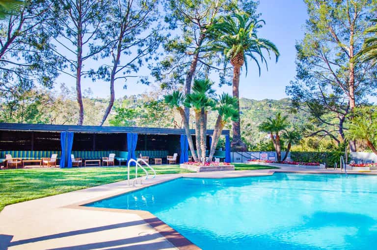 Resort pool surrounded by palm trees and tall pines with blue cabana structure and lounge chairs