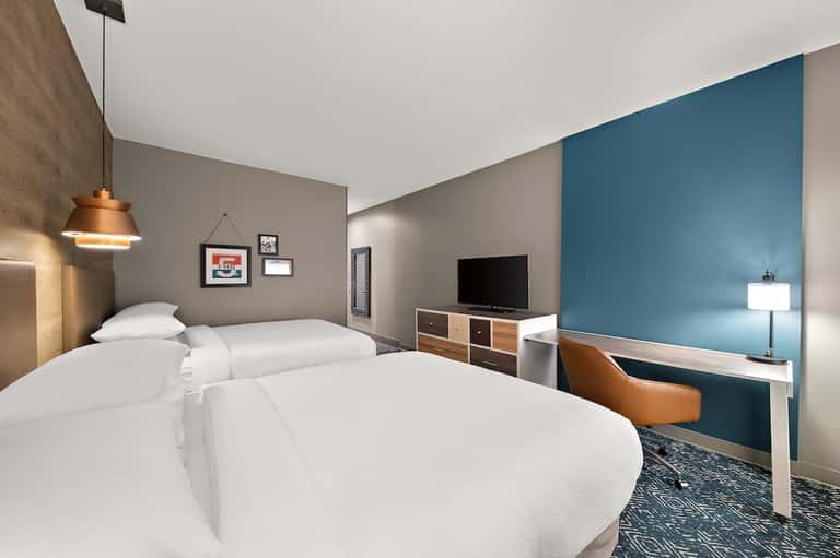 Contemporary hotel bedroom with two queen beds, blue accent wall, orange pendant light, desk, and neutral tones