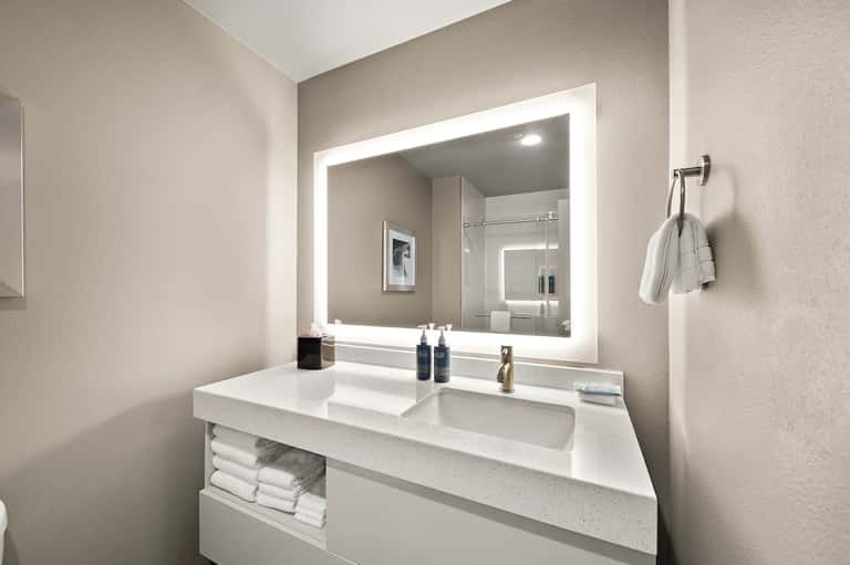 Modern bathroom vanity with white countertop, illuminated mirror, and shower visible