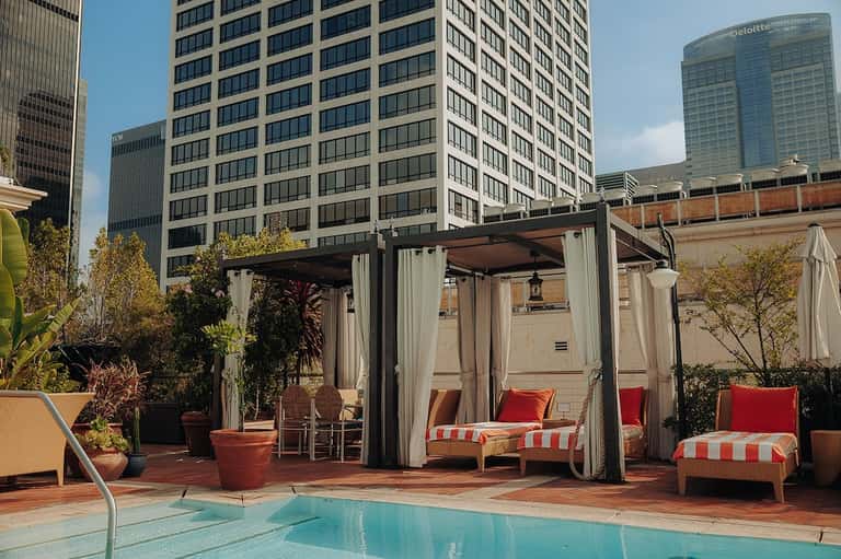 Urban rooftop pool area with lounge chairs, cabanas with red striped cushions, and city skyline backdrop