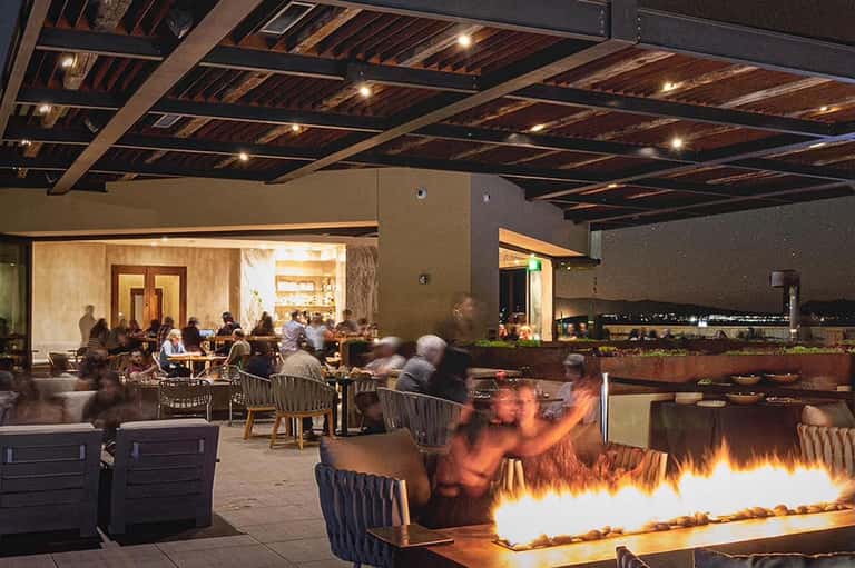 Rooftop dining area at night with fire pits, diners, wooden ceiling beams, and city lights in background